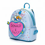 A light blue mini backpack featuring Donald Duck and Daisy Duck, with a large pink heart-shaped patch on the front that reads 'DONALD & DAISY' in black letters. The backpack is decorated with small multicolored hearts and has a blue top handle and zipper.